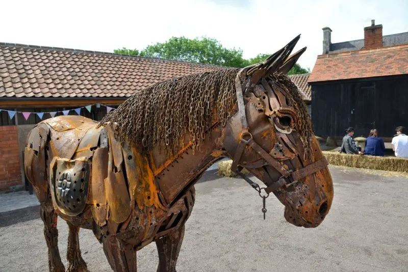 Large metal sculpture of a horse with saddle in a farmyard setting, with brick buildings and visitors in the background.