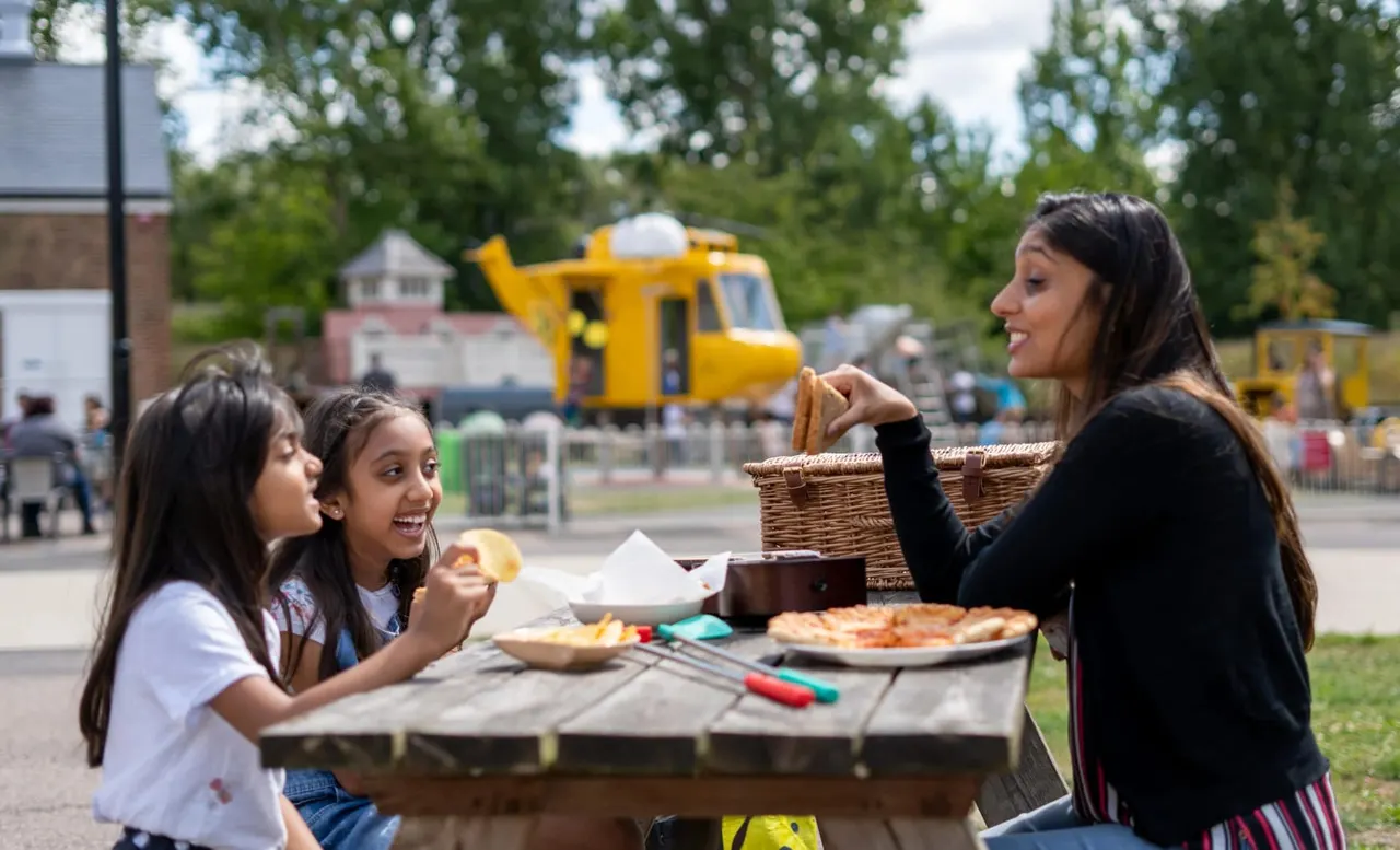An adult and two children having a picnic at an outdoor table, with a yellow RAF helicopter display visible in the background.