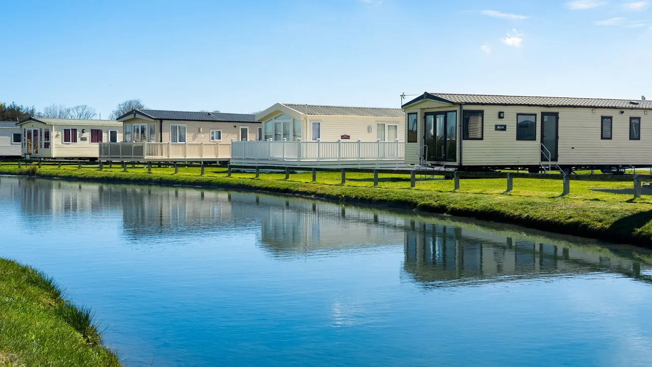 Row of static caravans on raised decking beside a calm waterway under clear blue sky.