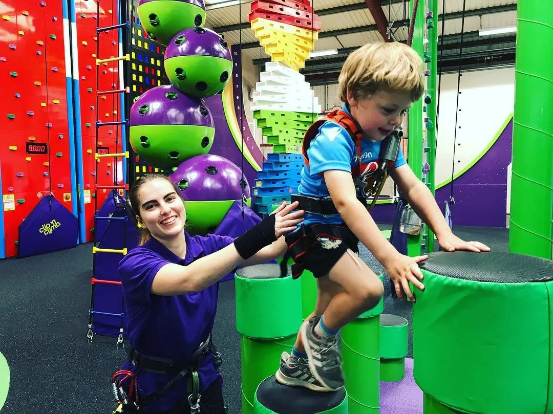 Staff member assists young boy navigating green climbing obstacles in colourful indoor adventure play facility.