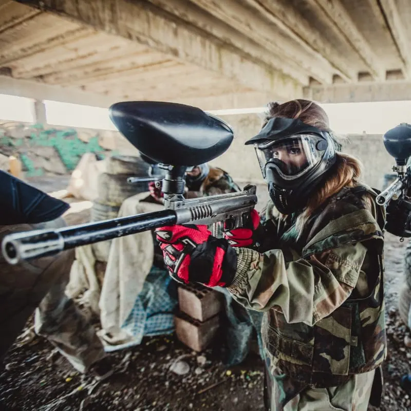 Player in protective mask and gear holding paintball gun at an outdoor paintball venue.