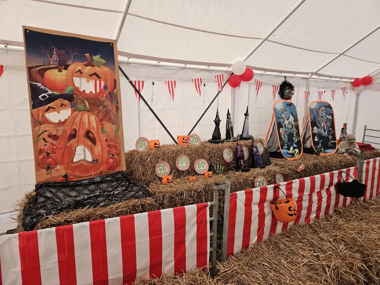 Halloween-themed indoor play area with hay bales, striped barriers, pumpkin game board, witch hats and decorations.