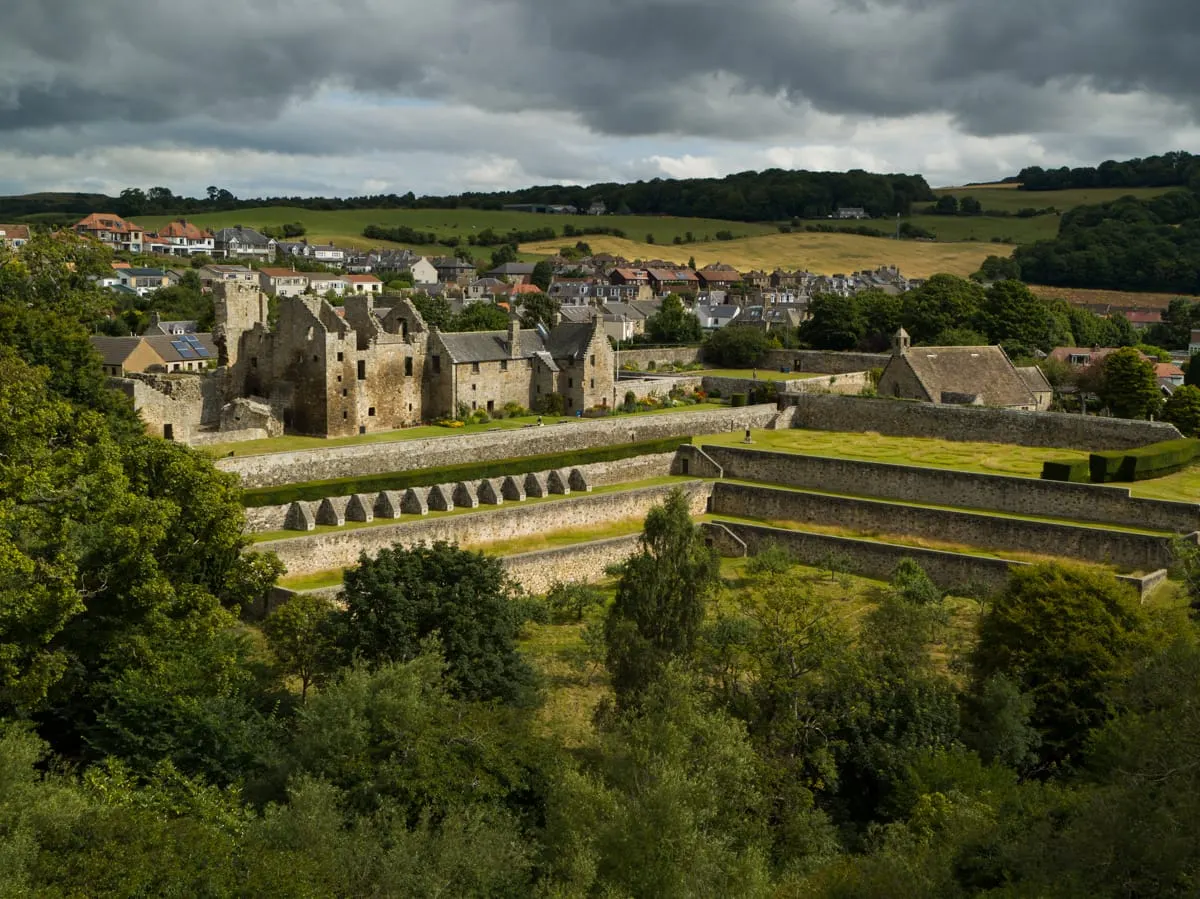 Medieval stone castle ruins with defensive walls and bastions overlooking a village nestled in rolling countryside under cloudy skies.