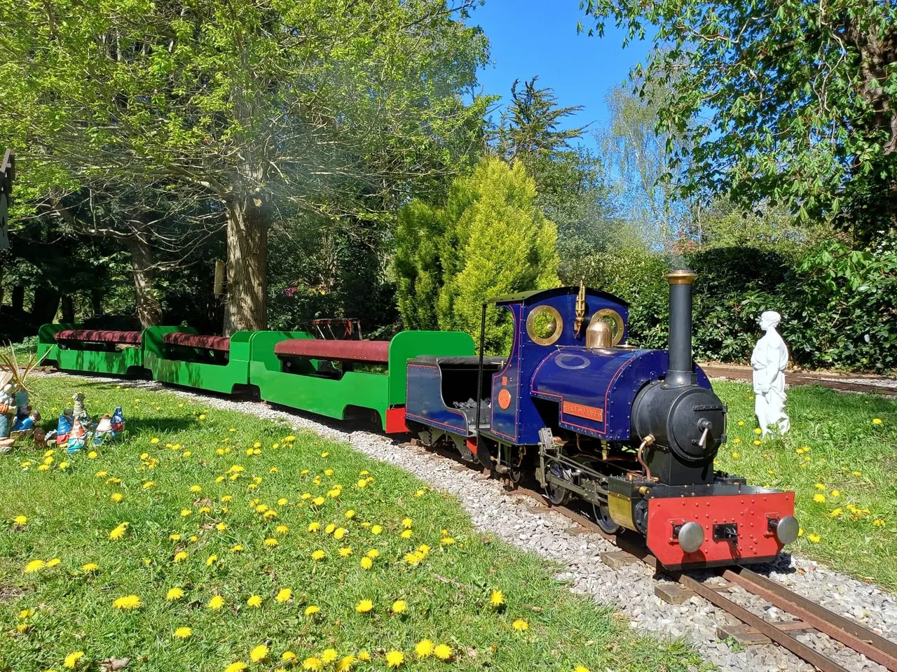 Miniature steam locomotive and green passenger carriages on a railway track through a garden with flowering grass and mature trees.