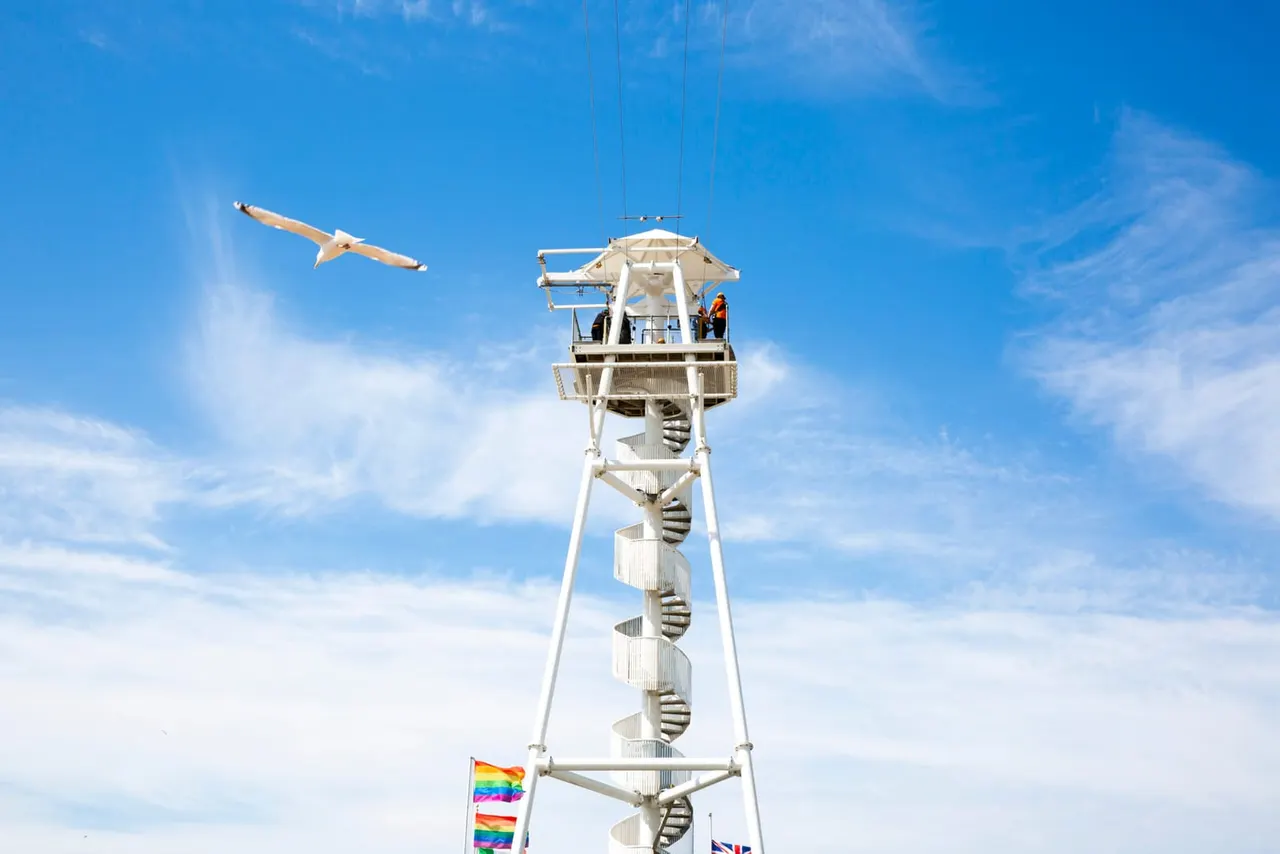 Two people descend a tall white spiral tower on a zip line against a clear blue sky.