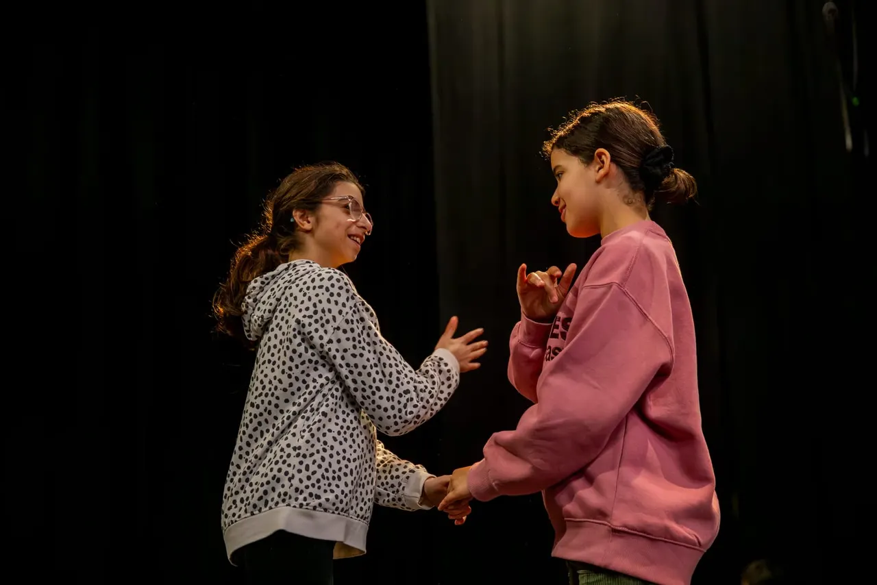 Two young people perform together on stage, facing each other with animated expressions and hand gestures against a dark background.