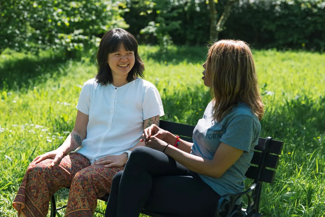 Two women sitting on a bench in a sunny green meadow, engaged in conversation.