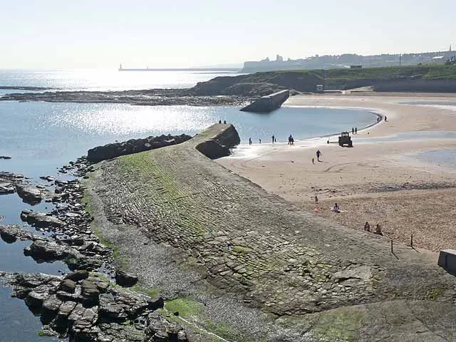 Sandy beach with rocky outcrops and shallow water, visitors exploring the shoreline and tidal pools.