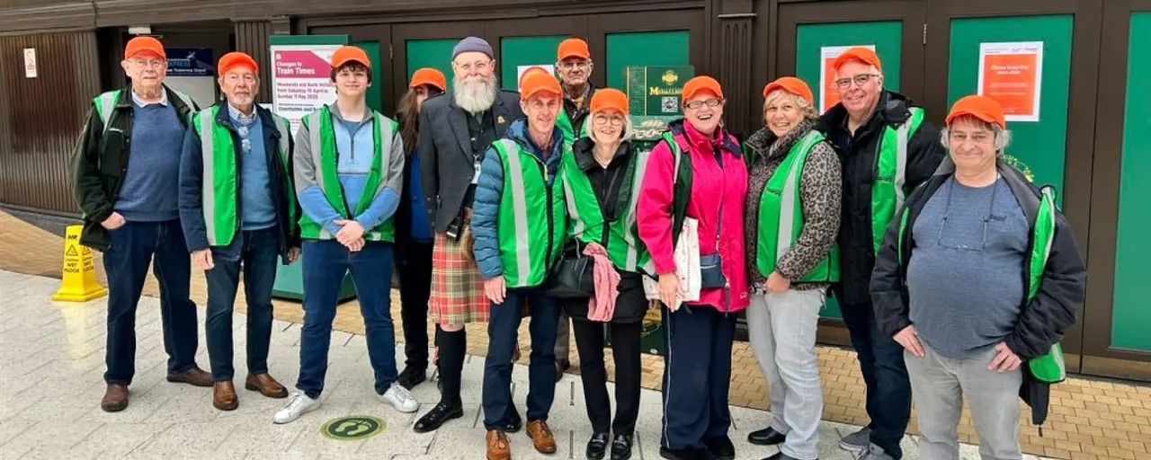A group of adult volunteers wearing orange caps and green high-visibility vests pose together outside a venue building.