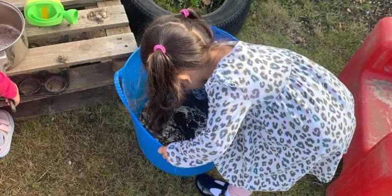 Young child leaning over a blue bucket outdoors in a garden activity area.