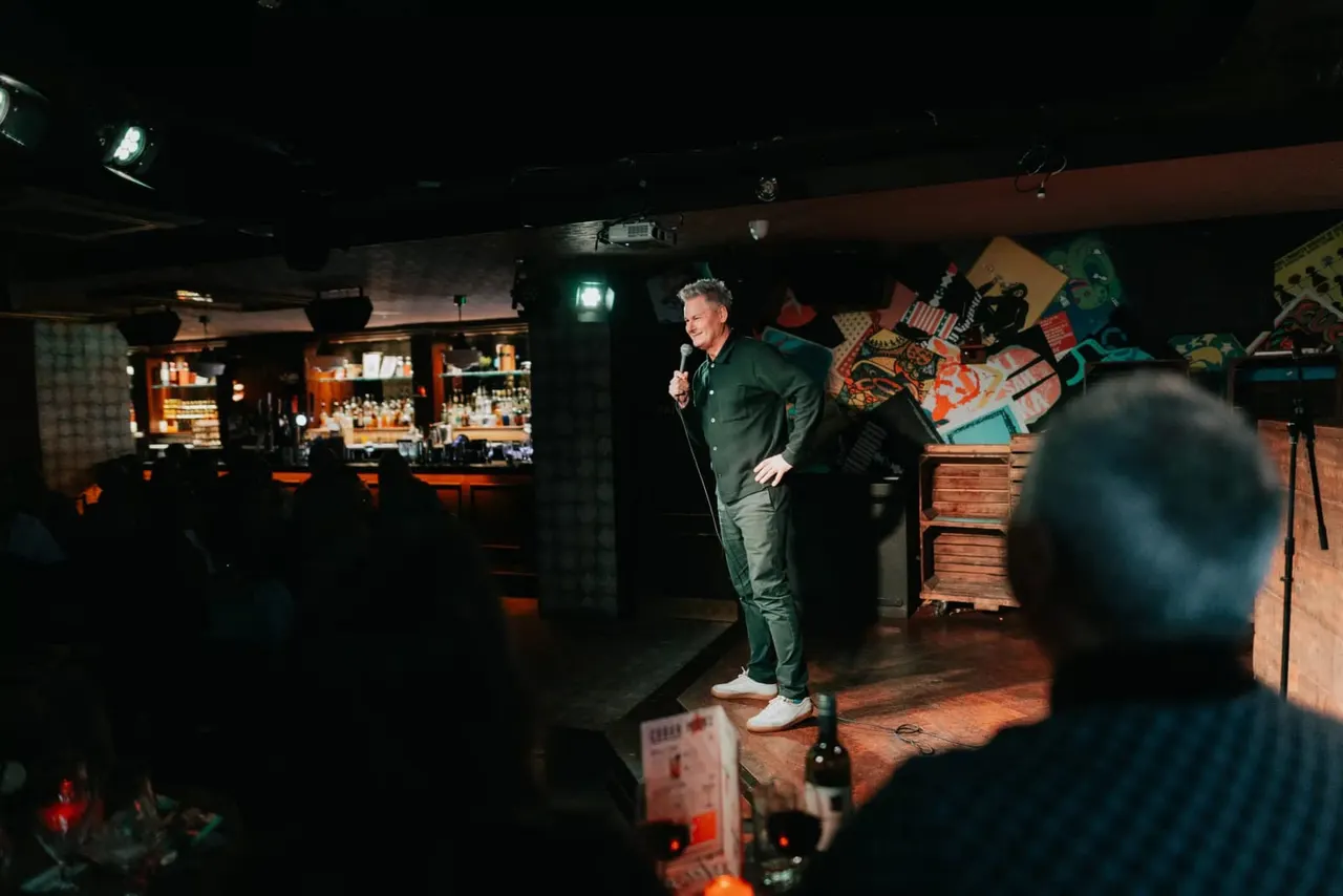 A man in a green suit performs stand-up comedy on stage with a microphone, facing an audience in a dimly lit venue with a bar visible in the background.