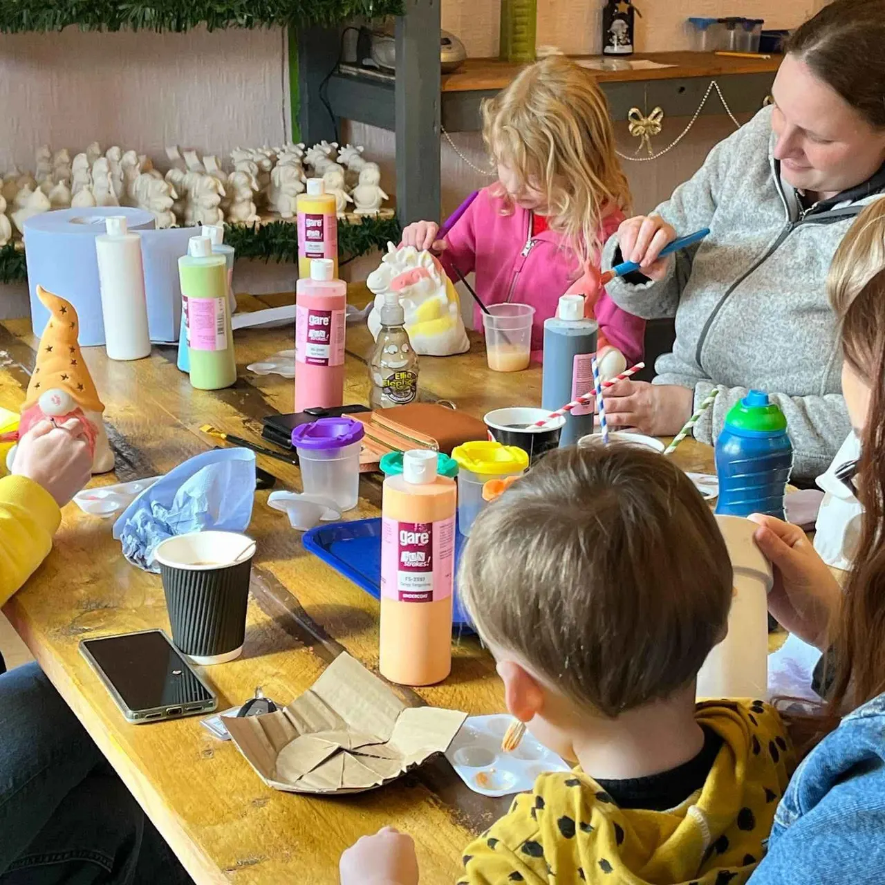 An adult and two young children doing a craft activity at a wooden table with paint, brushes, and decorative items.