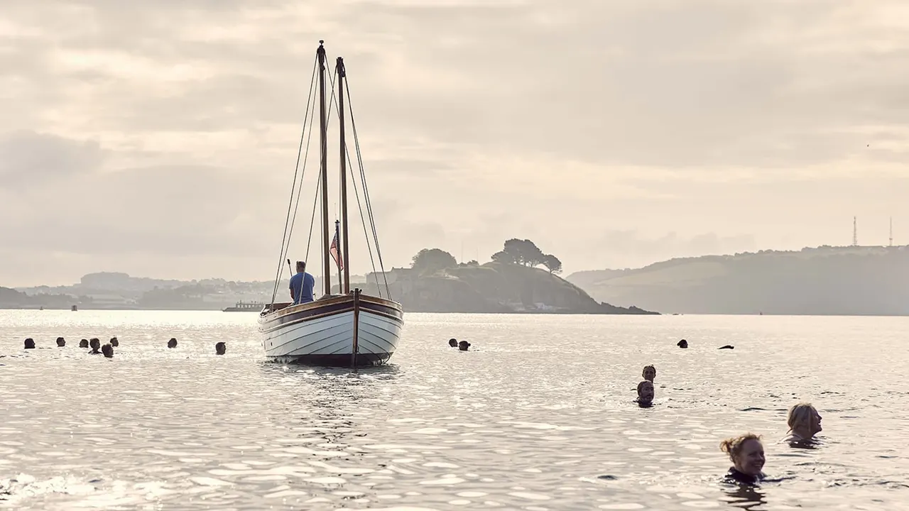 A sailing boat anchored in shallow coastal waters with swimmers and bathers in the sea around it, headland visible in the distance.