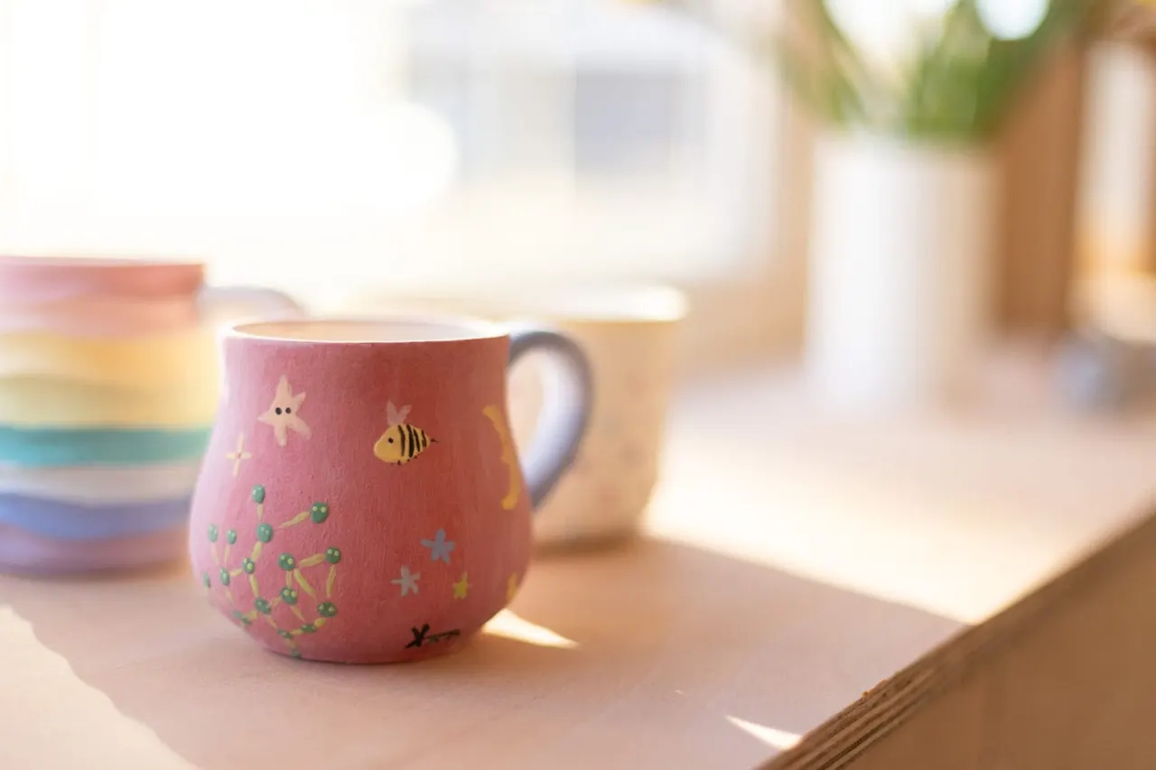 A pink ceramic mug decorated with stars, a bee, and floral designs sits on a wooden table beside colourful bowls.