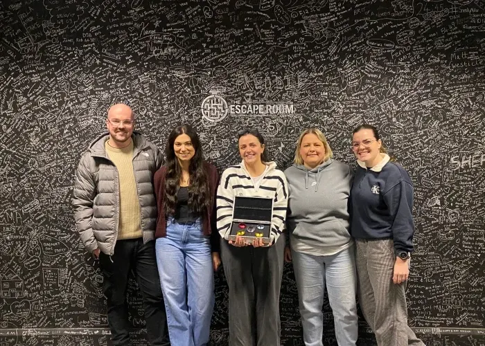 Five people standing together in front of a black wall covered with white doodles and the escape room logo.