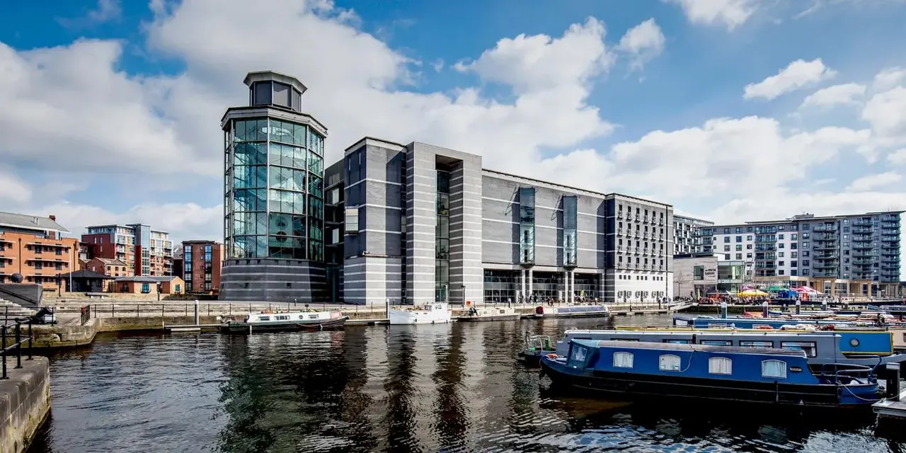 Modern waterfront building with glass tower beside moored boats on a canal, under blue sky with clouds.
