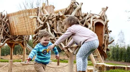 Young child and adult playing together at a wooden play structure made of branches in a rural outdoor setting.