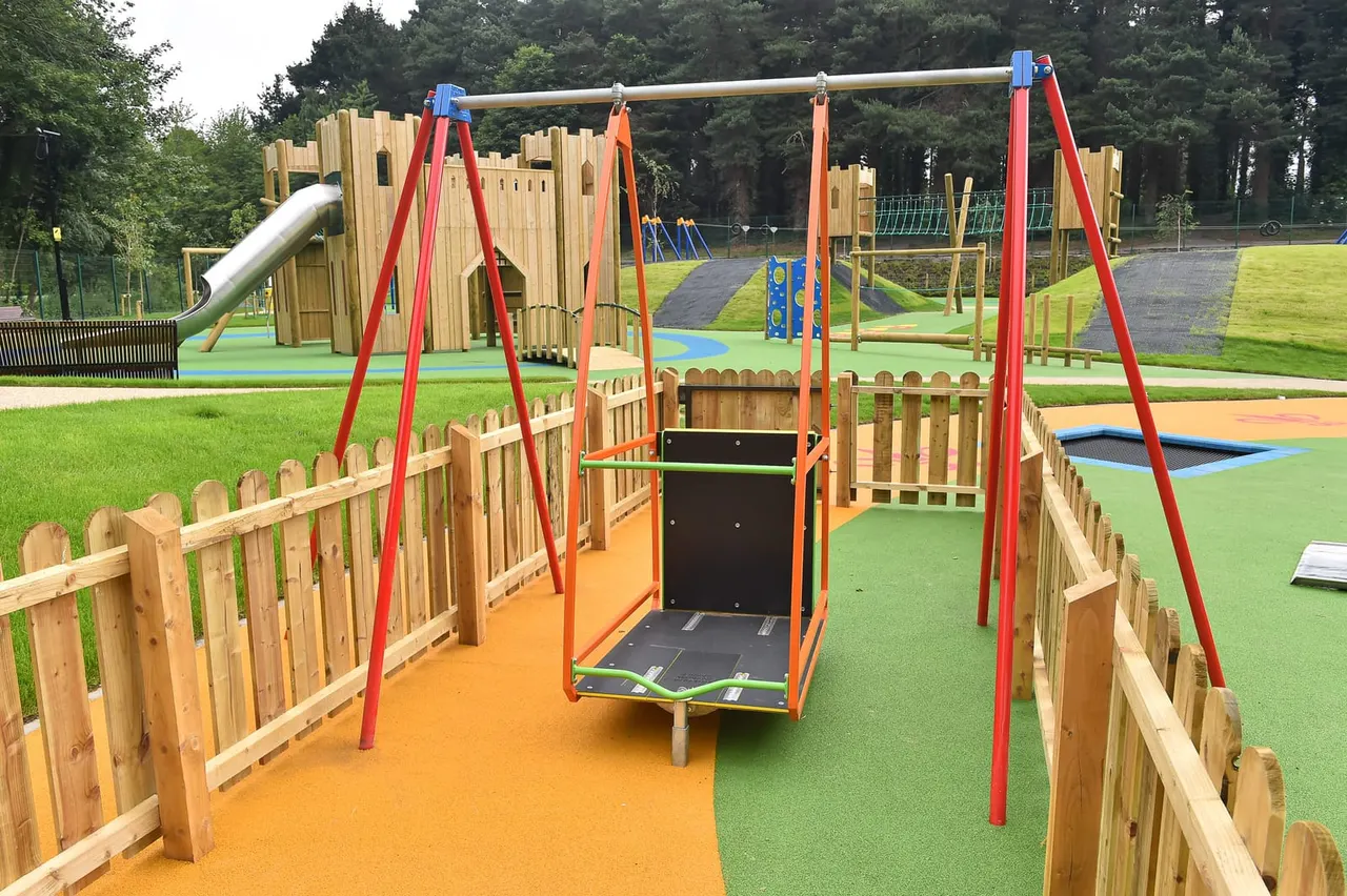 Outdoor play park with wooden fort structures, red metal swing frame with trampoline, and colourful surfacing surrounded by green grass and trees.