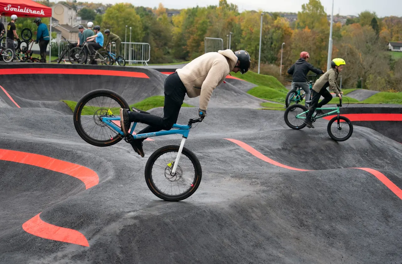 Rider performing a wheelie on a blue bike at a skate park whilst other cyclists ride on the curved concrete track.