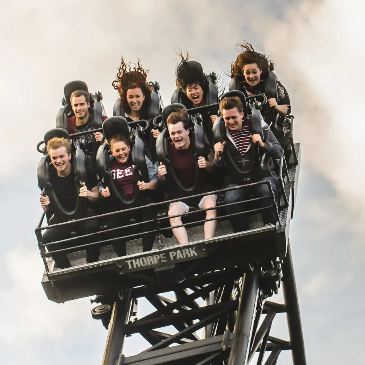 Group of young people on a roller coaster ride at the peak of a climb, smiling and reacting with excitement.