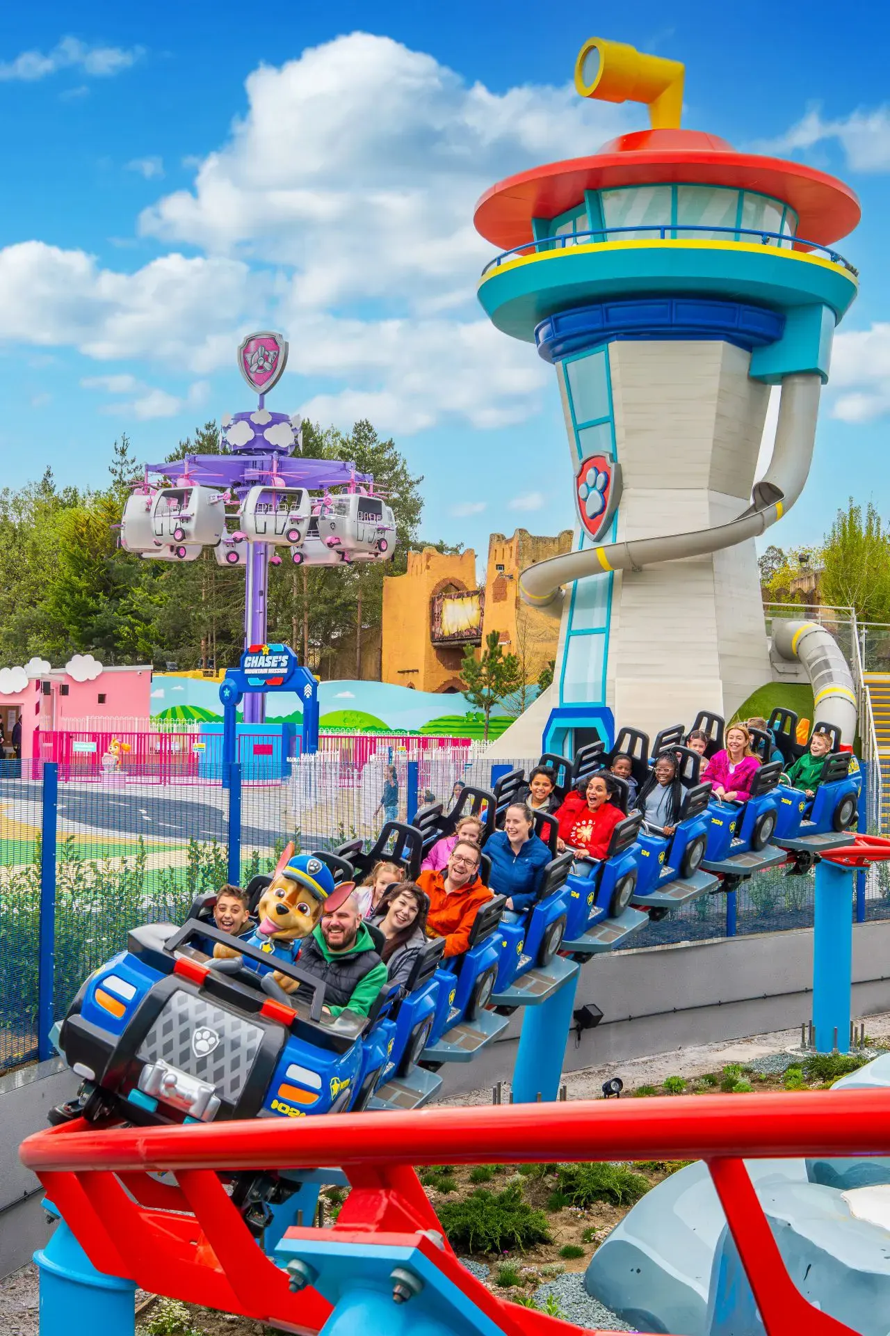Children and adults riding a blue coaster train past a colorful tower structure at an amusement park.