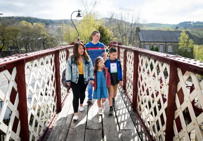 Family of four walking across a red and white lattice footbridge with rural countryside and stone buildings visible below.
