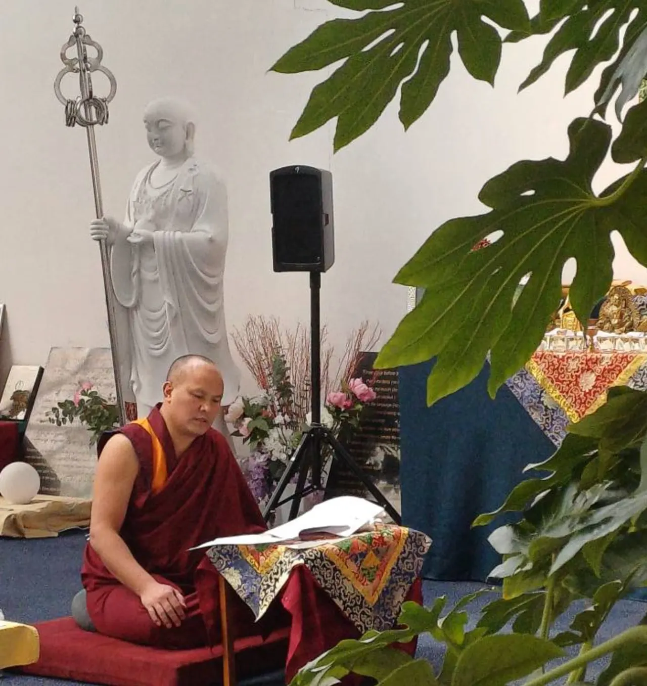 A Buddhist monk in maroon robes sits reading during a ceremony, with a statue and ceremonial items visible in the background.