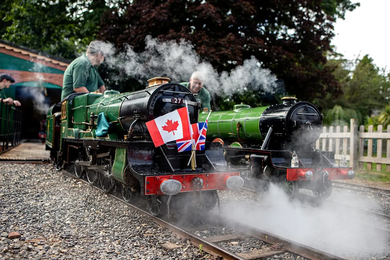 Steam locomotives numbered 27 and another engine steaming along a narrow-gauge railway track, with operators in green uniforms and international flags displayed.