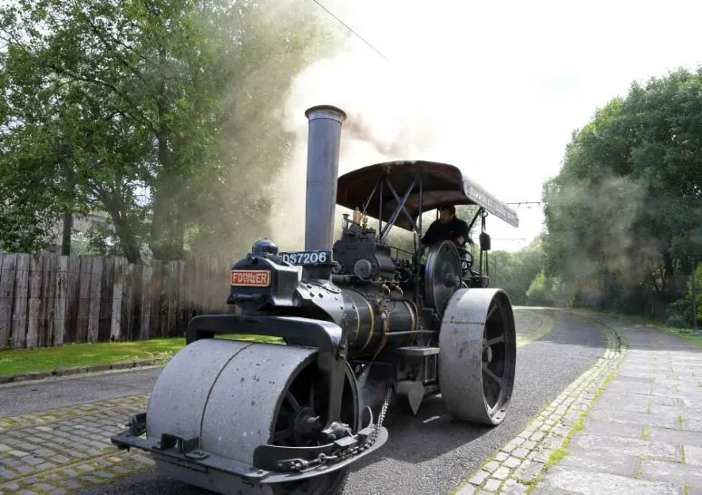 A vintage steam traction engine with tall smokestack travels along a tree-lined rural road, emitting steam and smoke.