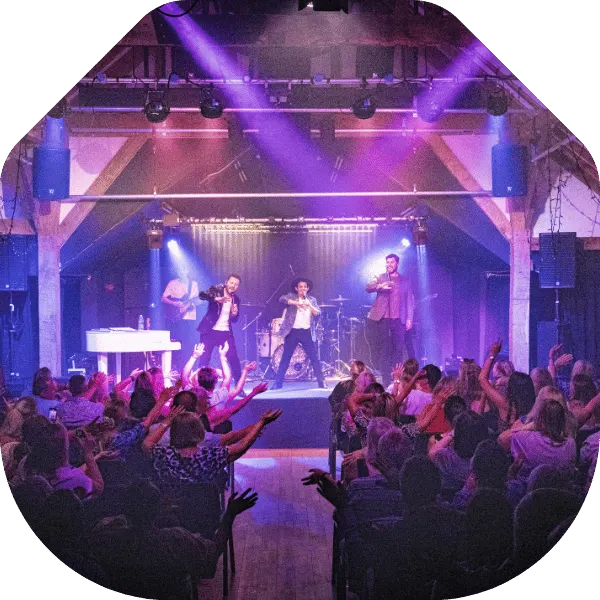 Band performing on stage with purple lighting as audience members dance and watch in an indoor venue.