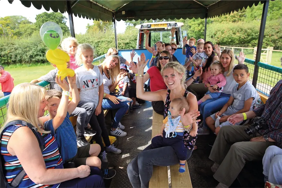 A large group of adults and children sitting together on a ride vehicle under a green canopy, waving and smiling at the camera with a tractor visible behind them.
