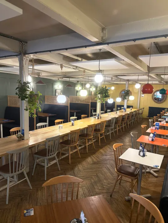 Modern café interior with long wooden communal table, individual dining tables, hanging pendant lights, and wooden chairs.