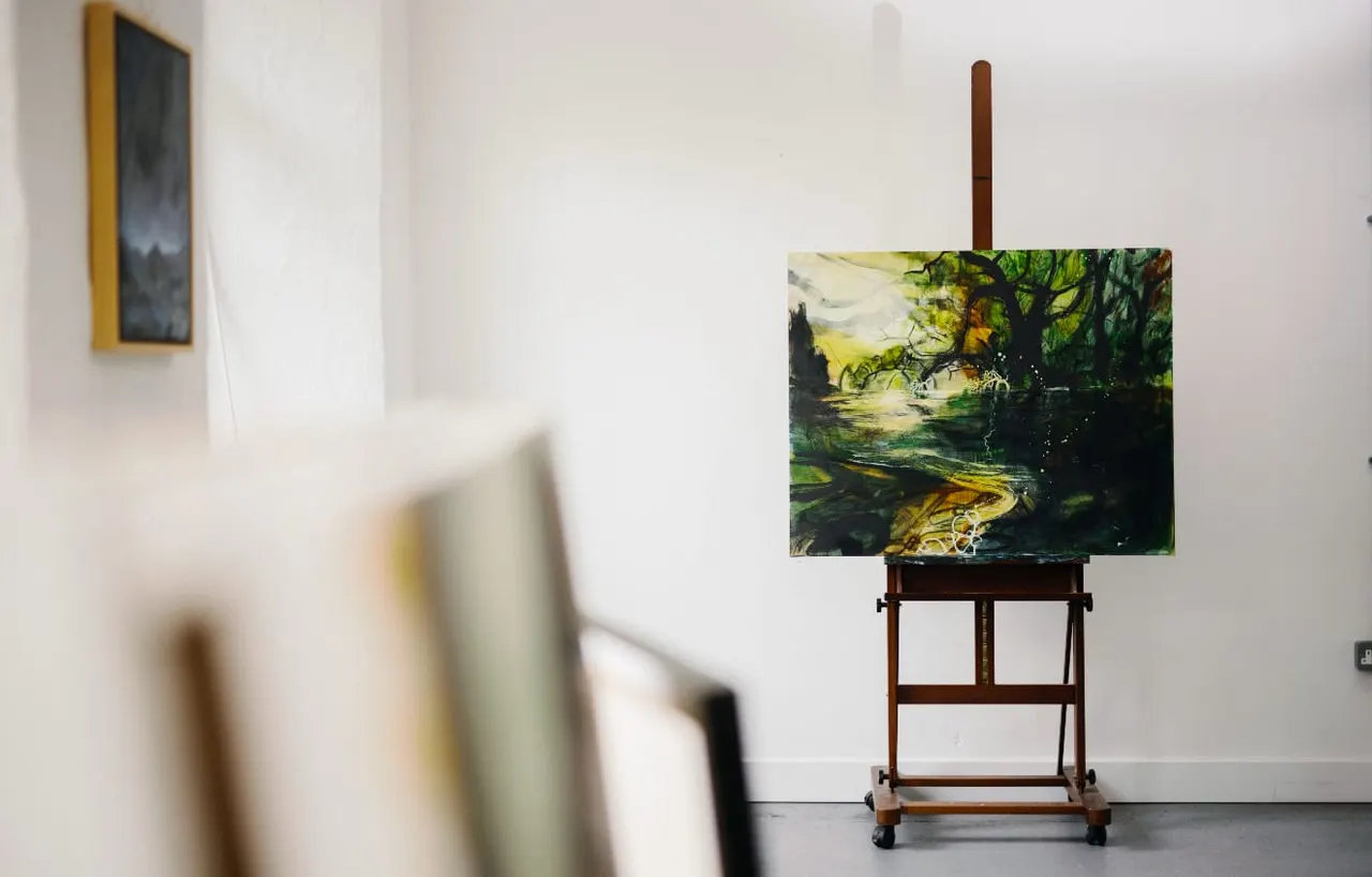 Painting of a river landscape with trees displayed on an easel in a gallery space with white walls.