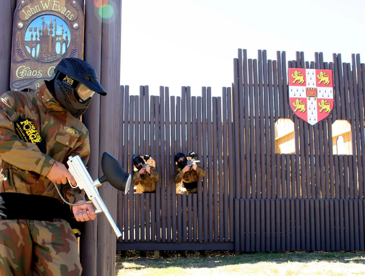 A paintball player in camouflage gear and cap holds a paintball marker whilst standing near a wooden palisade fortress wall with heraldic shields.