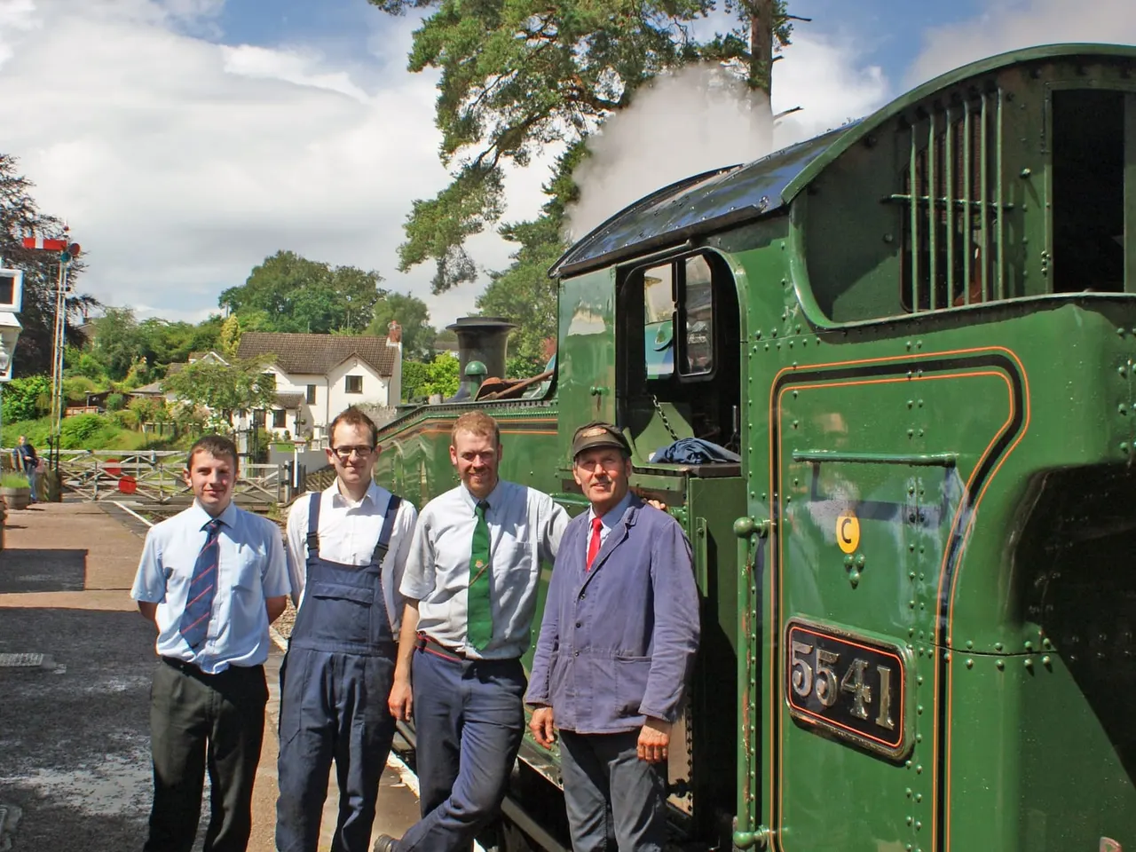 Four male staff members in period uniforms stand beside a green steam locomotive at a heritage railway station.