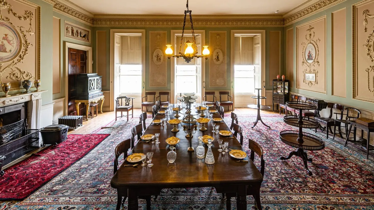 Ornate Georgian dining room with long wooden table set for formal dinner, gold-trimmed walls, ornamental ceiling, and period furniture.