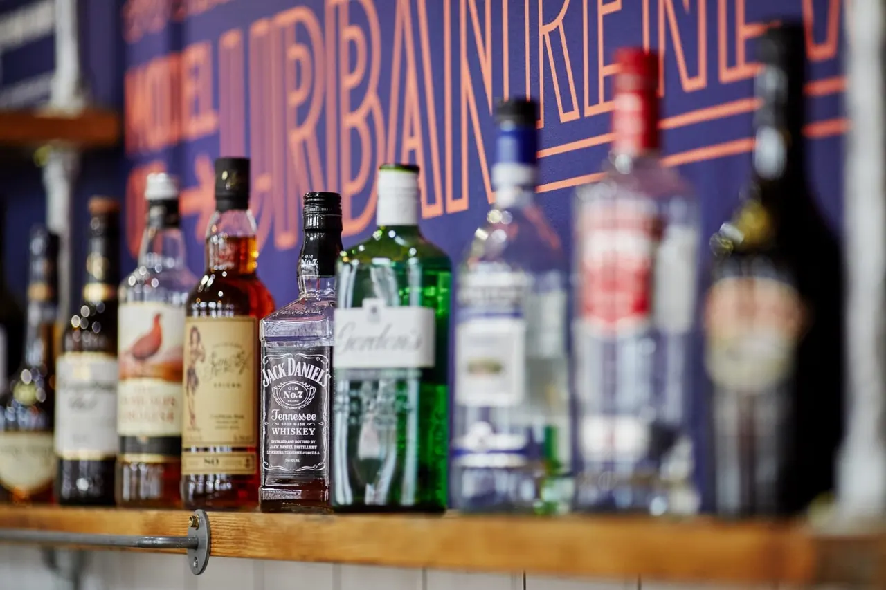 Array of spirits and wine bottles displayed on a wooden bar shelf with a neon sign blurred in background.