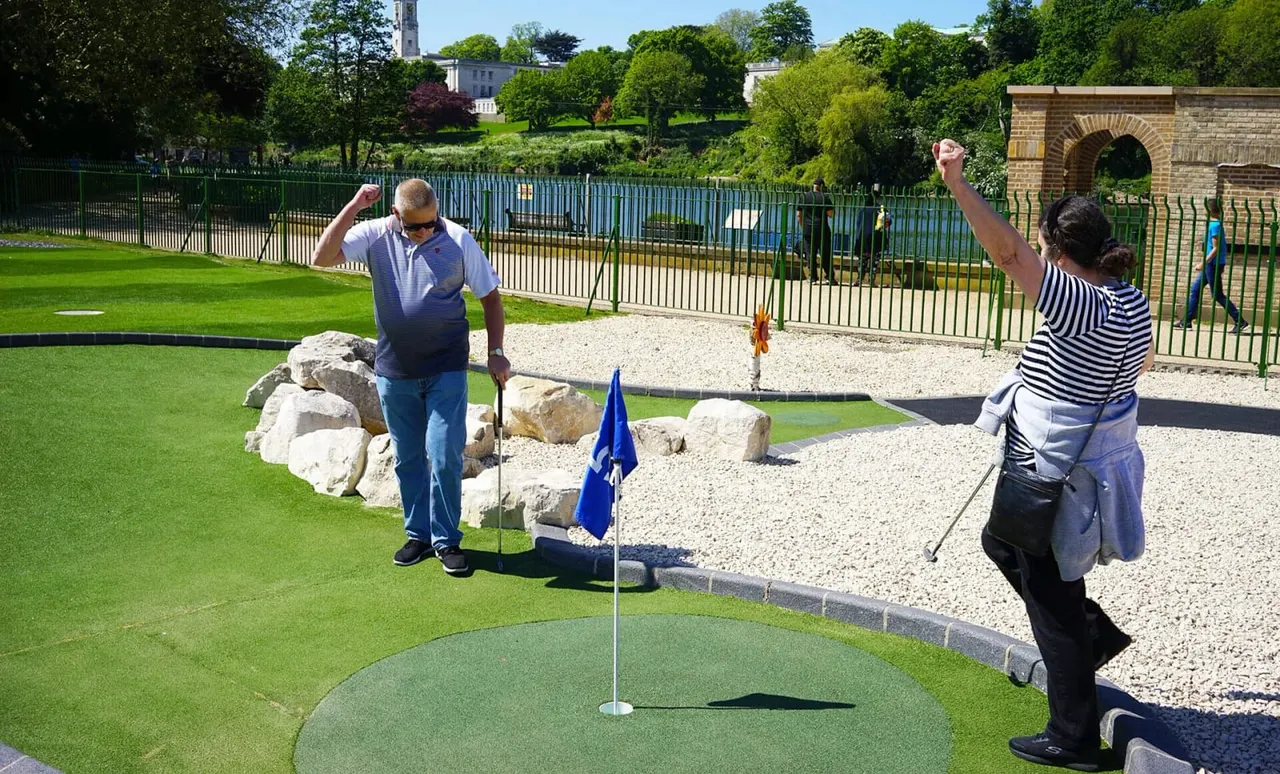 Two adults playing miniature golf on a bright sunny day, with park grounds, fencing and historic buildings visible in the background.