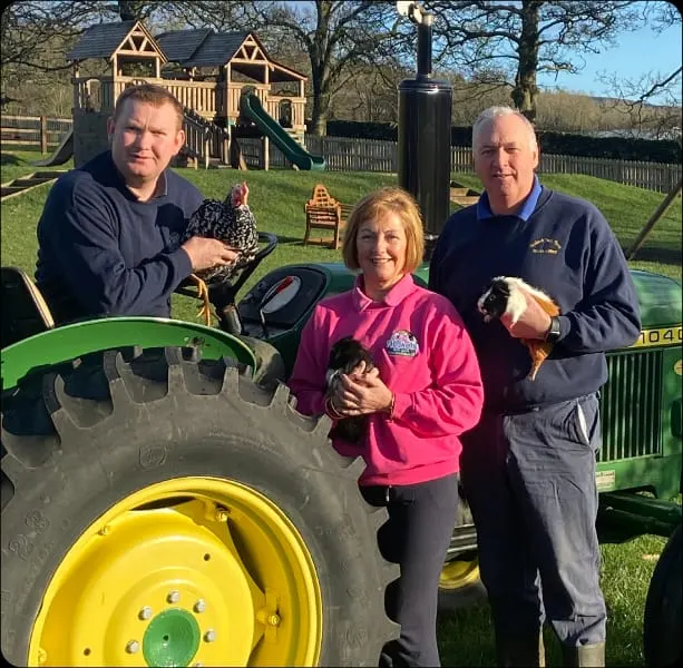 Three adults holding farm birds next to a large green farm vehicle tyre on a sunny day.