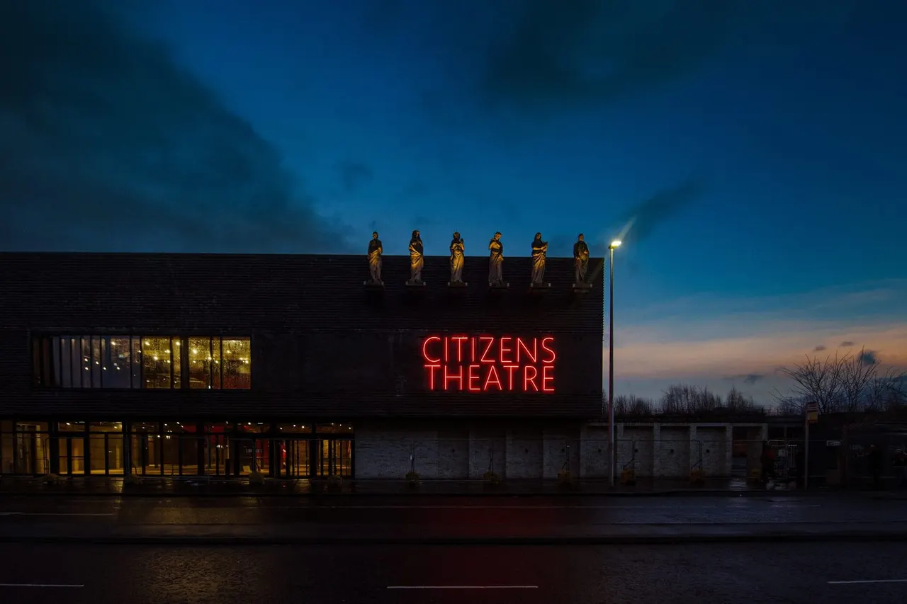Modern theatre building with red neon sign lit at dusk, statues on roofline, and lit windows below against twilight sky.