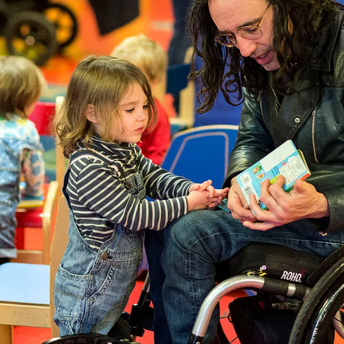 Adult in wheelchair reading a picture book with a young child in striped top in a colourful indoor play space.