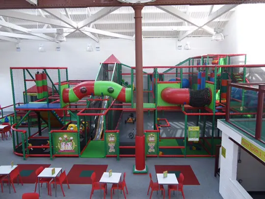 Colourful indoor play structure with slides, climbing frames and tunnels in bright green and red, with seating area visible in foreground.