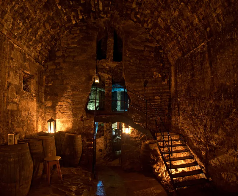 Historic stone cellar with wooden barrels, lantern lighting, and metal staircase leading upwards.