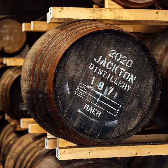 Wooden barrels stacked in a distillery, with a labelled cask bearing "2020 Sackton Distillery 1871" in the foreground.