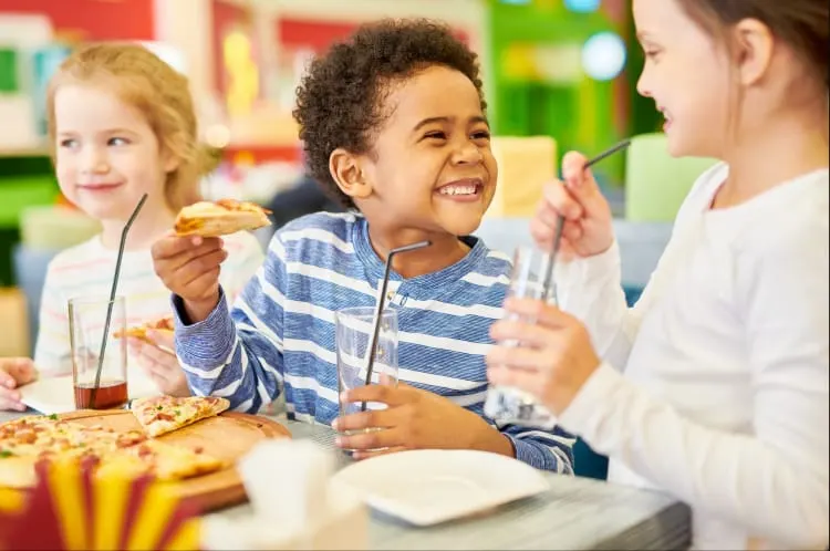 Three young children eating pizza and drinking soft drinks together at a colourful play centre.