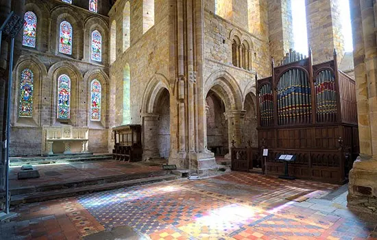 Stone church interior with stained glass windows, ornate organ, and patterned floor bathed in coloured light.