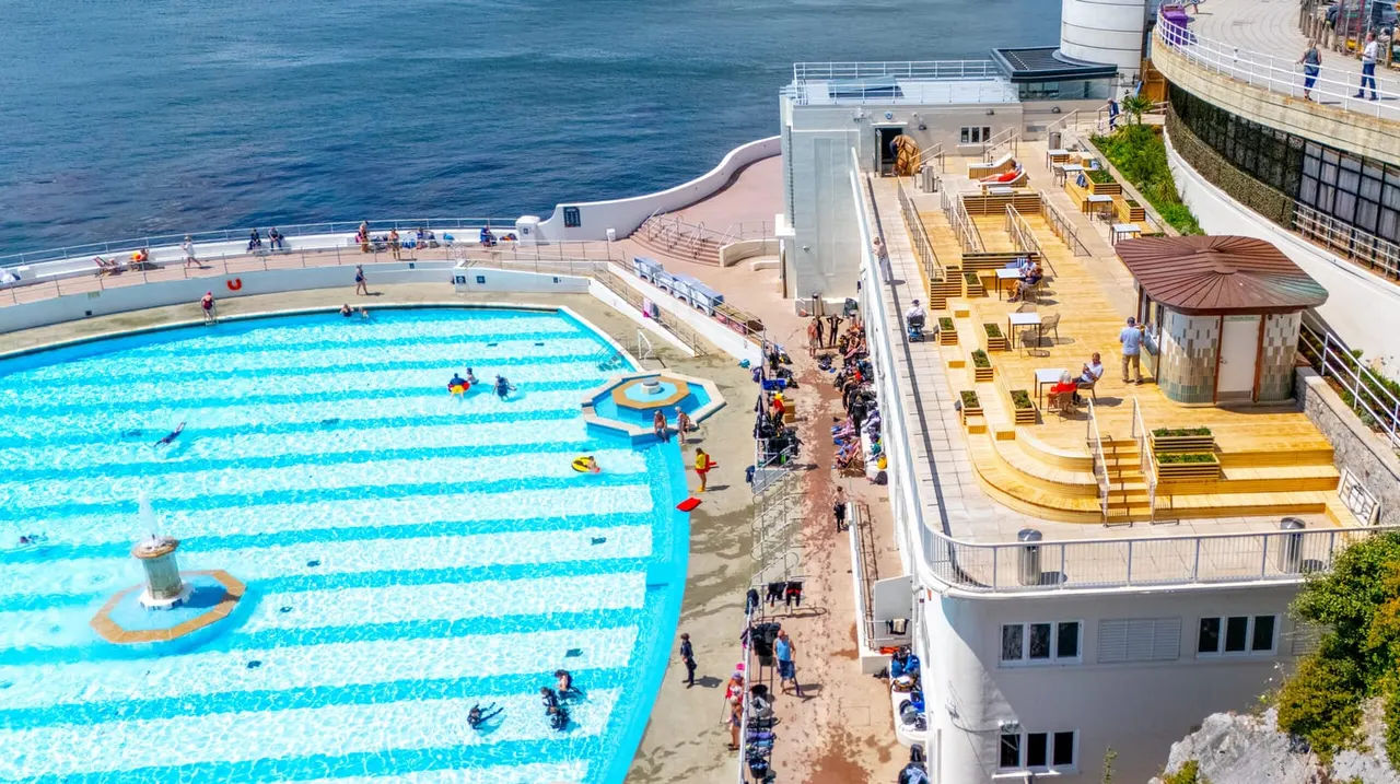 Aerial view of a seaside lido with a large swimming pool, tiered seating areas, and visitors enjoying the facilities by the sea.