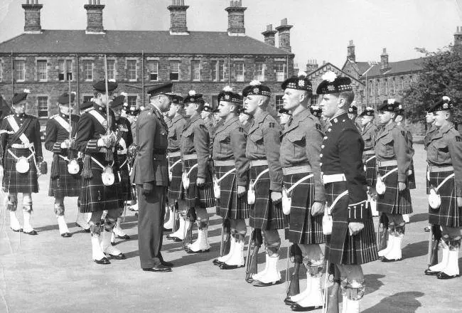 Military personnel in formal dress uniforms and kilts assembled in a courtyard parade, with historic stone buildings behind them.