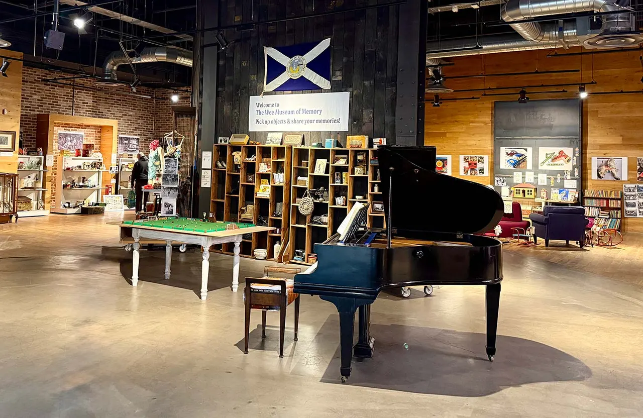 Interior of The Wee Museum of Memory showing a grand piano, pool table, wooden shelving units filled with objects, and a visitor browsing displays in an industrial-style space.