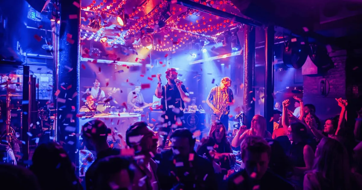 Live band performing on stage to a dancing crowd under colourful neon lights and hanging lanterns in a nightclub venue.